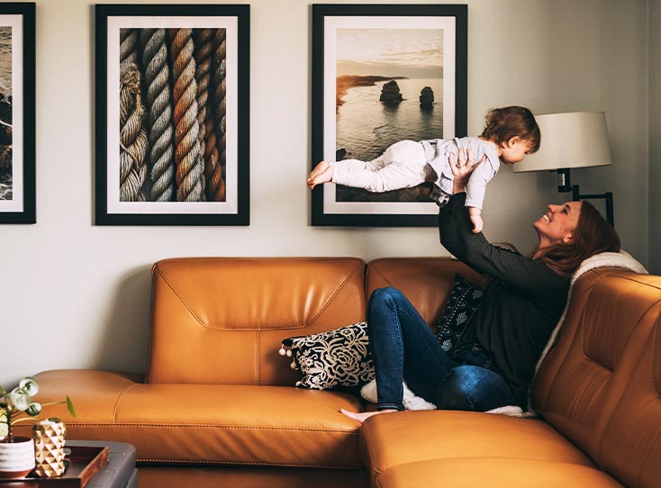 Woman on orange leather couch holding up young child in front of a three-frame gallery wall